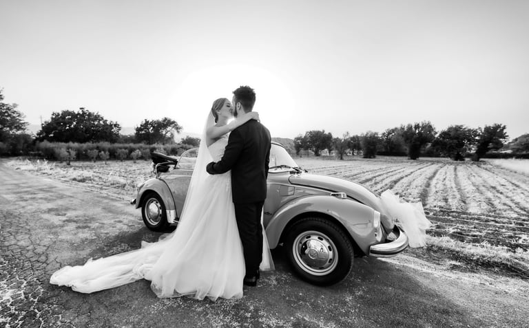 Sposi che si abbracciano accanto a un’auto d’epoca decorata, in un paesaggio di campagna al tramonto