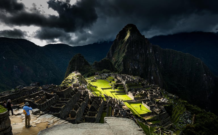 This image shows Machu Picchu with dark stormy skies contrasting sunlight that illuminates the ancient Inca ruins in Peru.
