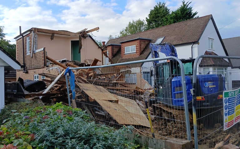A blue excavator demolishing a residential house behind a construction safety fence.