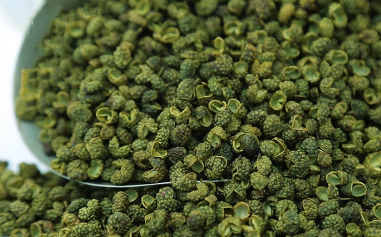 A metal scoop overflowing with dried green Sichuan peppercorns, showcasing their unique husks and texture.