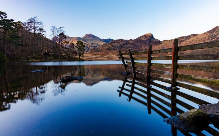Rustic wooden fence reflecting in the calm blue water of a lake with scenic mountain peaks.