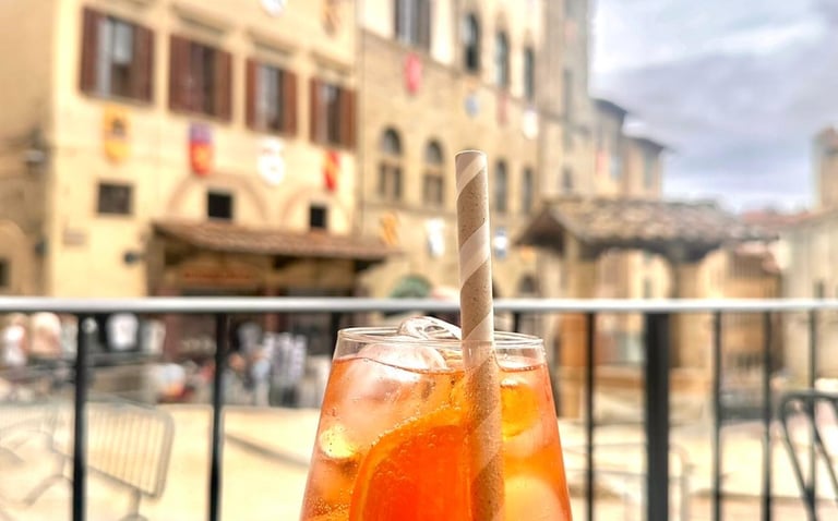 Refreshing orange Aperol Spritz cocktail with a paper straw at an outdoor cafe in a historic Italian piazza.