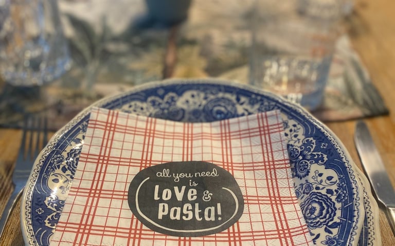 Rustic dinner table setting featuring blue patterned plates and a napkin reading All You Need Is Love and Pasta.