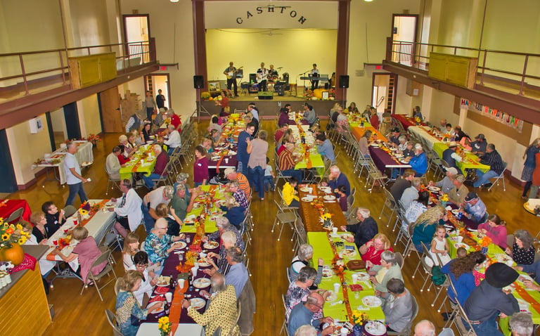 a large group of people sitting at tables with food eating together with a band on the stage