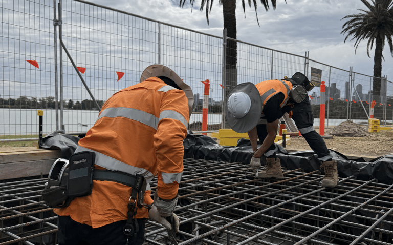 Construction workers in high-visibility safety gear install steel reinforcement mesh for a concrete foundation.
