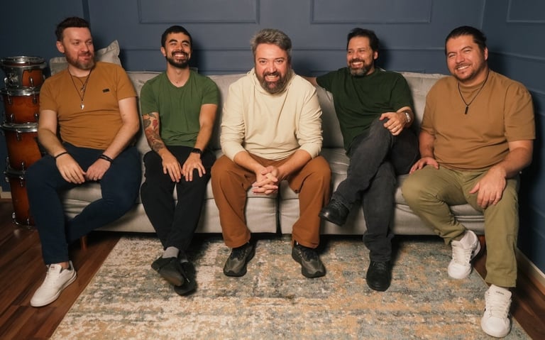 A smiling five-member rock band sitting on a grey sofa in a studio with drums visible in the background.