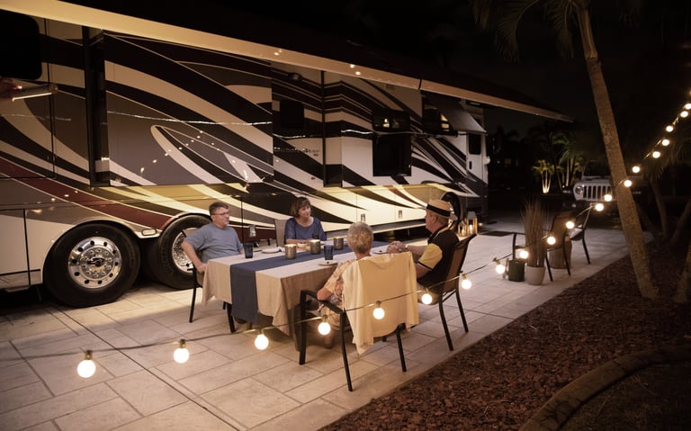 Friends enjoy outdoor dining by a luxury motorhome RV at night with festive string lights.