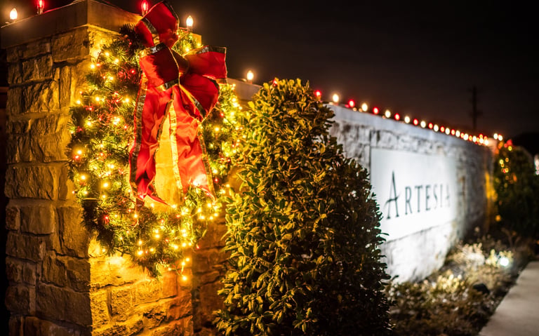 A festive Christmas wreath with a red bow and glowing lights on a stone wall entrance to HOA.