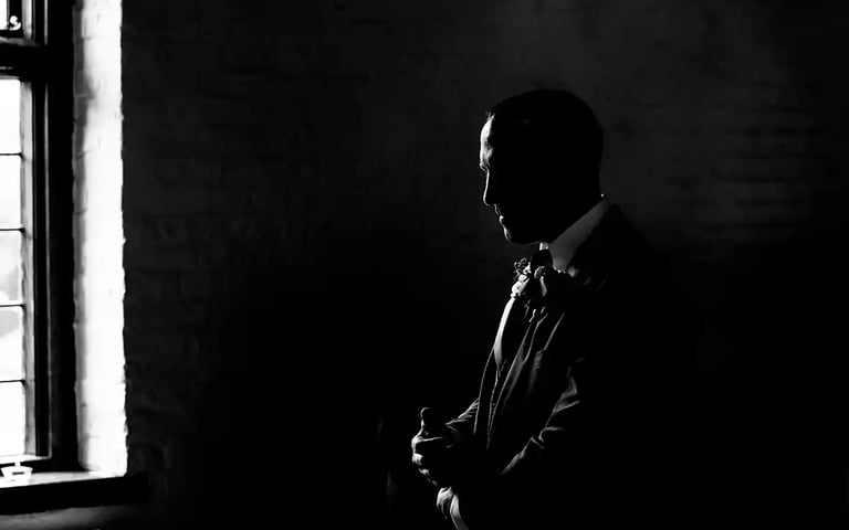 Black and white silhouette of a groom in a tuxedo standing by a window before a wedding ceremony.