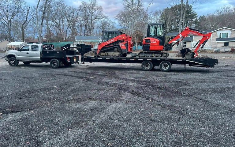 A silver flatbed truck towing a Kubota skid steer and mini excavator on a gooseneck trailer.
