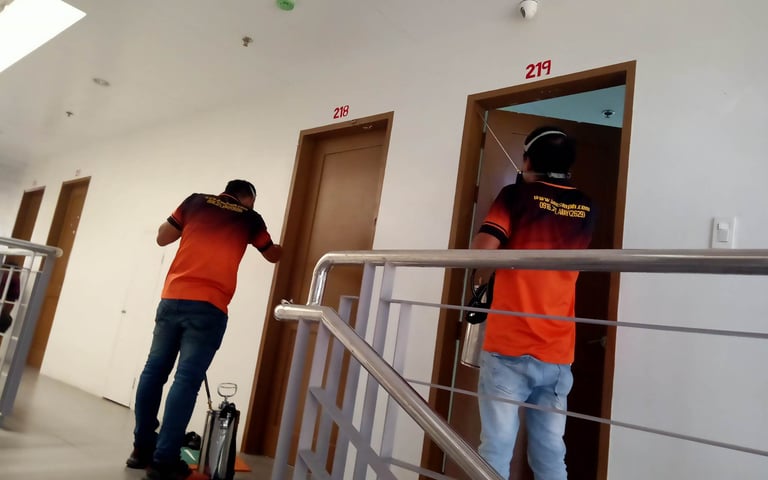 Professional pest control workers in orange uniforms spraying insecticide near apartment doors in a hallway.