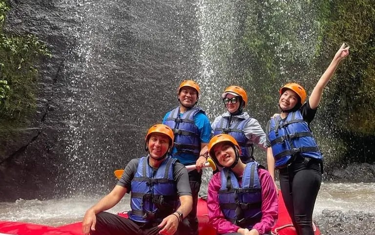 A group of friends in helmets and life jackets posing in a red white-water raft near a waterfall.