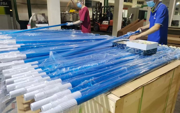 Factory workers packing blue telescopic pool cleaning poles into protective plastic sleeves for shipping.