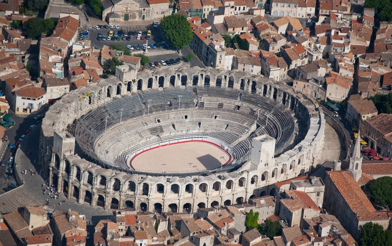 High-angle view of the ancient Roman amphitheater in Arles, France, surrounded by historic city buildings under a bright sky.