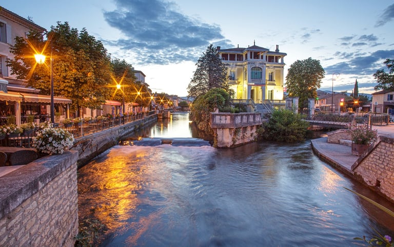 Evening view of L'Isle-sur-la-Sorgue canal in Provence with illuminated riverside cafes and a grand historic building.