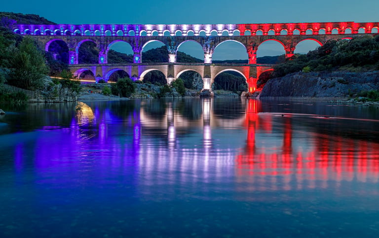 Pont du Gard Roman aqueduct illuminated with blue, white, and red lights reflected in the Gardon River at night.
