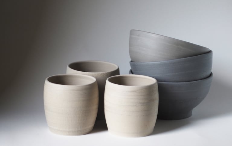 Trys smėlio spavos žiesti rankų darbo neglazūruoti puodeliai, šalia jų trys pilki bliūdukai