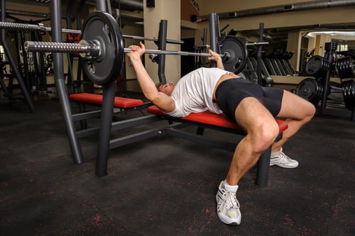 A muscular man performing a barbell bench press on a weight bench in a commercial gym setting.