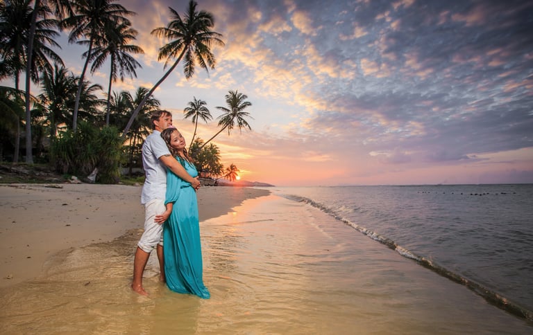 A couple embracing on a tropical beach at sunset, with palm trees and the ocean in the background