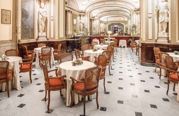 Elegant hotel dining room with marble floors, white statues, and round tables with orange chairs.