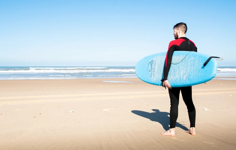 En homme en combinaison de surf, avec une planche bleu sur une plage