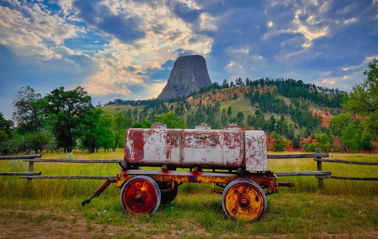 Rustic vintage water wagon in a field with Devils Tower National Monument in the background under a dramatic sky.