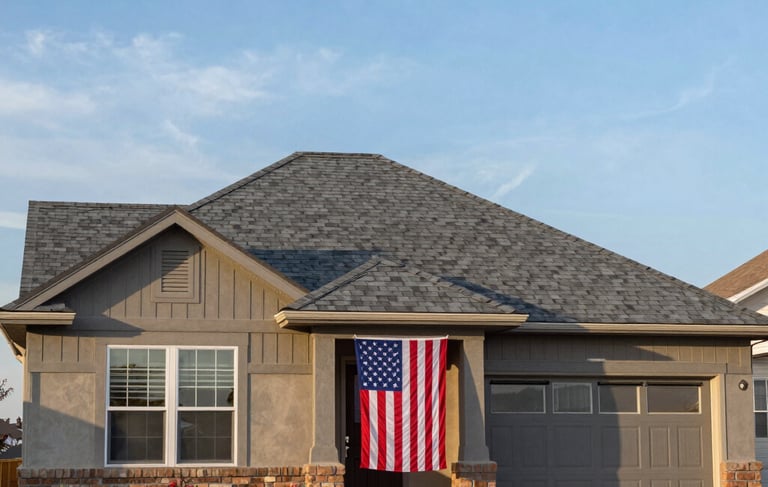 A clean, eye-level photograph of a residential house in Colorado Springs with a brand new asphalt shingle roof in a medium gray color. The house has a well-maintained lawn and a North American / US flag proudly displayed by the front door. The sky is a bright blue with soft morning light, and the overall feel is one of quality craftsmanship and patriotic pride.