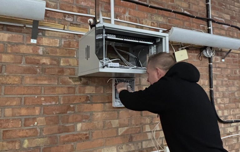 An IT technician installs network cabling and server equipment in a wall-mounted rack on a brick wall.
