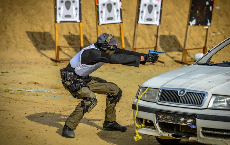 Tactical shooter in training gear aims a blue handgun from behind a car at an outdoor firing range.