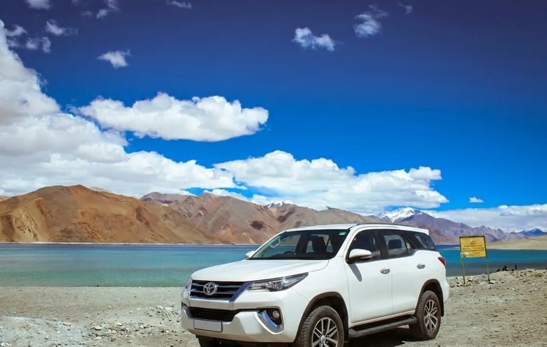 White Toyota Fortuner SUV parked by the scenic Pangong Lake in Ladakh under a blue sky.