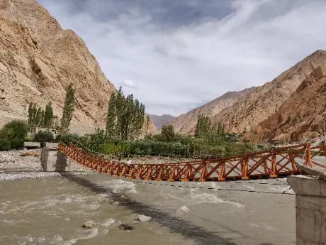 Orange suspension footbridge crossing a river in a rocky mountain valley landscape.