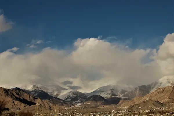 Panoramic view of a Himalayan mountain range with snow-capped peaks and scattered clouds over a town in Ladakh.