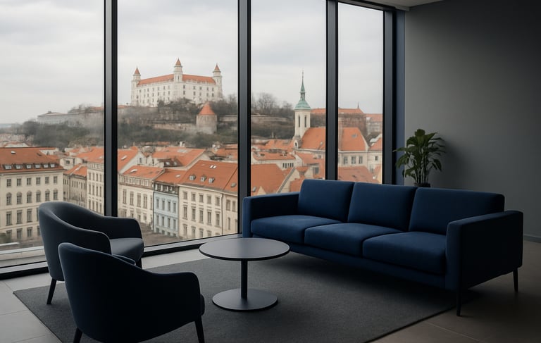 A crisp, high-end commercial photography shot of a modern office lounge in Bratislava, Slovakia. Through a floor-to-ceiling glass window, clean Central European city architecture is visible under soft overcast light. The interior features sleek furniture in midnight blue and muted grey textiles. A professional atmosphere reflecting synergy and reliability, with natural daylight and a minimalist composition.