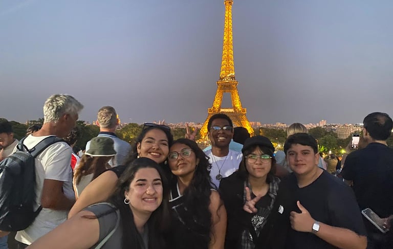 A imagem mostra um grupo de estudantes em frente à Torre Eiffel iluminada, em Paris.