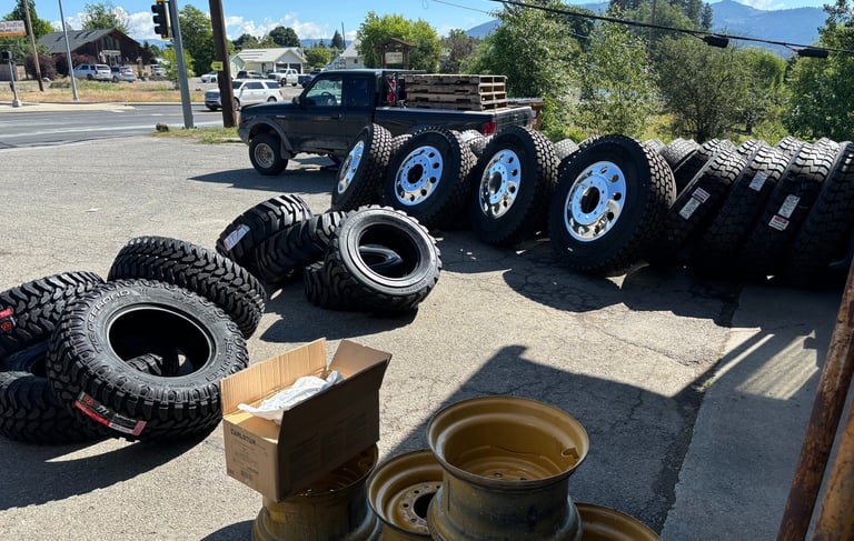 A collection of heavy-duty truck tires and chrome rims displayed on asphalt at a tire service shop.