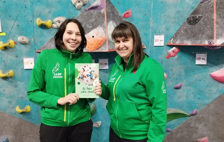 two women in green jackets standing next to a climbing wall holding a book