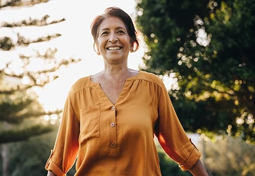 A smiling senior woman in an orange blouse enjoying a sunny day outdoors at a park.