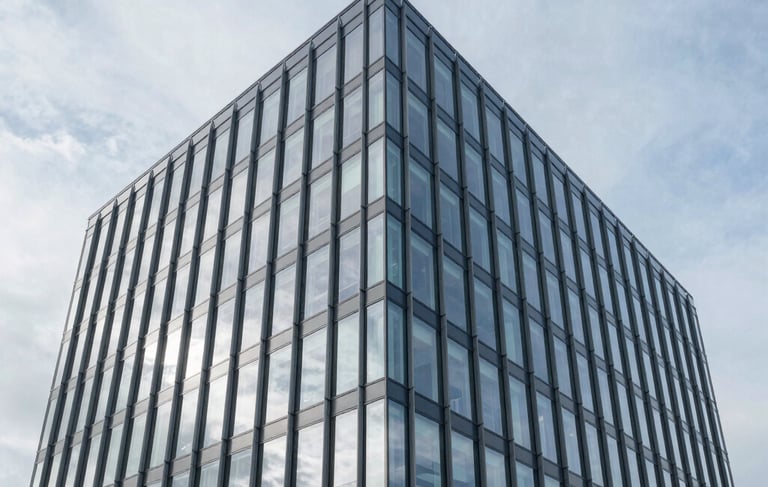 A sharp, wide-angle architectural photograph of a modern commercial office building in Ilford, United Kingdom. The image features a clean glass facade reflecting a light grey and medium blue sky, conveying a sense of transparency and modern professionalism. The lighting is bright and clear, emphasizing reliability.