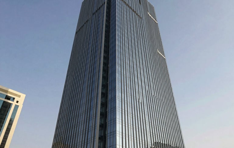 A professional wide-angle architectural photograph of a sleek glass office tower in the Ajman Free Zone. The building reflects a clear sky, featuring sophisticated shades of dark navy and silver. The scene is set in a modern Middle Eastern / Gulf commercial district, conveying a sense of authority and global reach through clean lines and bright, natural sunlight.