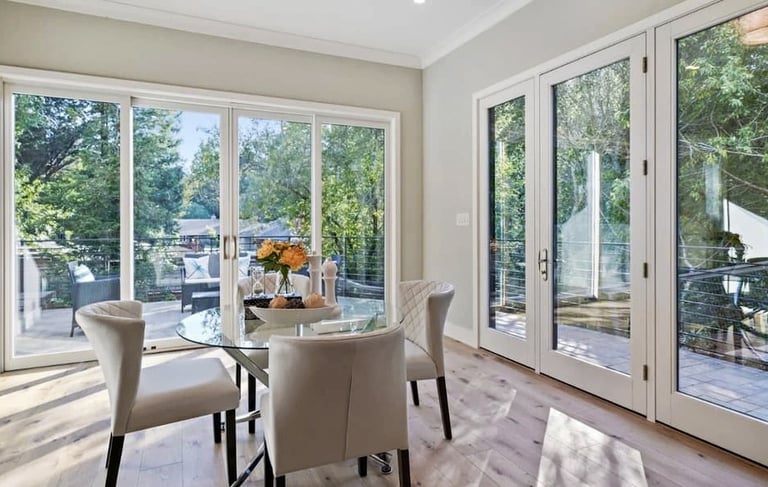 Dining area with a glass table and large windows overlooking an outdoor space.
