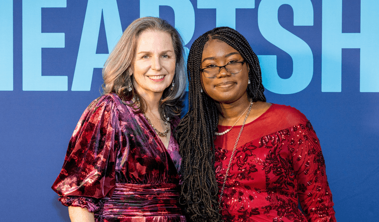 Two women in formal red and purple dresses smiling at a Heartshare event with blue background.
