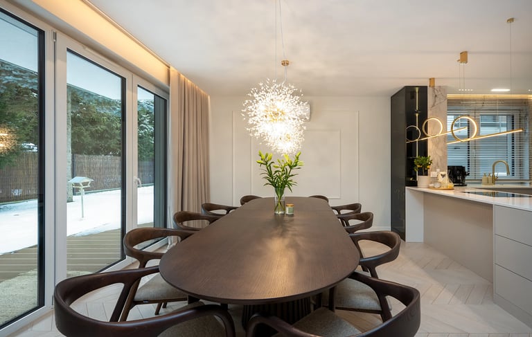 Nowoczesna jadalnia z długim stołem, krzesłami, lampą sufitową i kuchnią po prawej.