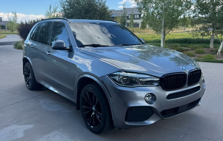 A silver BMW X5 luxury SUV with black rims parked on a driveway under a blue sky.