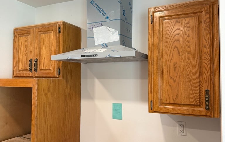 A stainless steel range hood being installed between oak kitchen cabinets during a home remodel.
