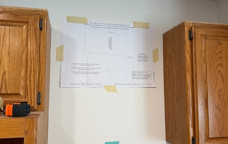 Paper mounting template for range hood installation taped to a kitchen wall between wood cabinets.