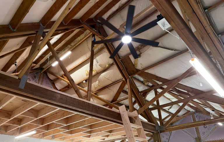 Exposed wood roof trusses in a garage attic with a black ceiling fan and LED shop lights.