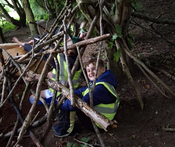 Cub Scout sitting inside a den made from sticks during a forest survival activity.