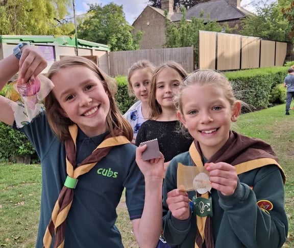 Two smiling Cub Scouts proudly showing materials from an outdoor activity, with other Cubs behind th