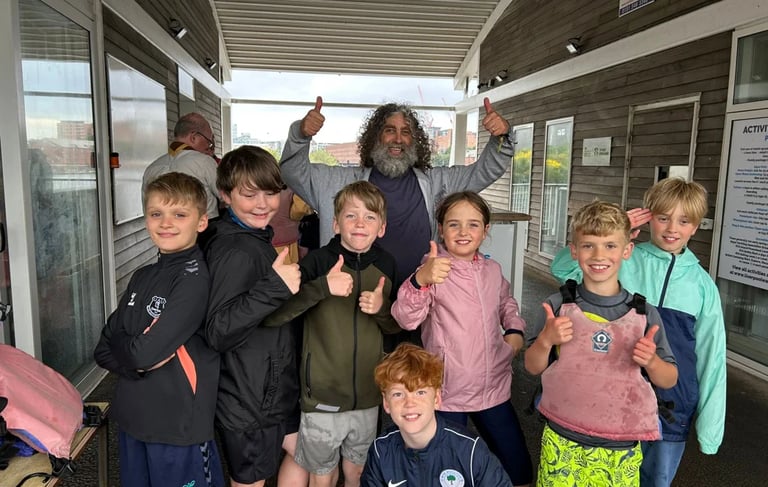 Group of Cub Scouts giving thumbs up with a scout leader under a shelter at a waterfront venue.