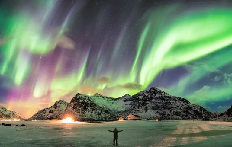 A person stands under the vibrant green and purple aurora borealis over snowy mountains in Norway.